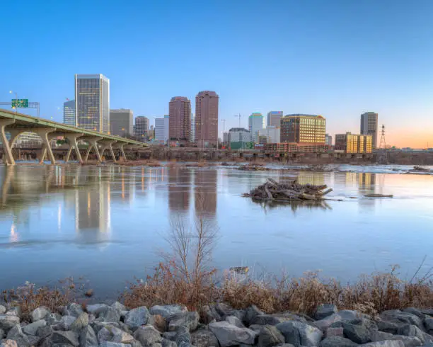 The city skyline of Richmond Virginia is visible above the James River at sunrise.  A cool morning blue sky reveals the lights of skyscrapers at dawn.