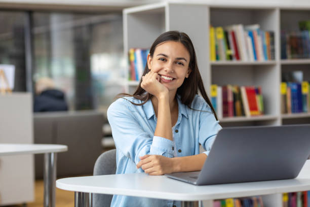 Beautiful female student or female employee sitting at table in the university library or office with laptop looking at the camera Remote work or education concept stock photo