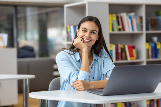 linda estudante ou funcionária sentada à mesa na biblioteca ou escritório da universidade com laptop olhando para a câmera trabalho remoto ou conceito de educação - professor da faculdade - fotografias e filmes do acervo