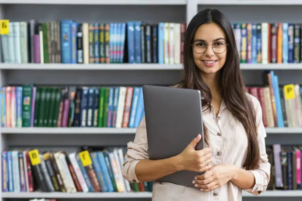 Beautiful female student with laptop smiling on the background of the library bookshelves. Learning and education online concept