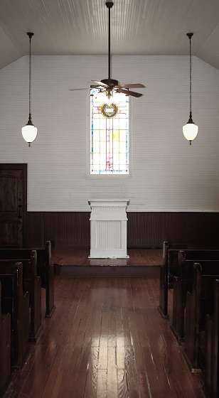 looking down the aisle at an old church altar or pulpit in wood, white with a stained glass window behind, ceiling fan and light above and two hanging lights on either side, wooden floor and the ends of the pews in dark mahogany wood