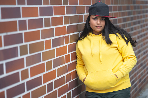 Smiling latino girl with a yellow hoodie in front of a brick wall with hands in the pockets and a baseball hat