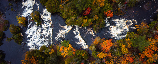 Incredible panoramic travel aerial look down photograph of Bond Falls Scenic Site including the upper and lower waterfalls and rapids in autumn with fall colored foliage on the treetops below.