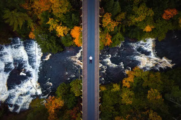 Photo of Beautiful autumn look down travel aerial of a man laying down on the abandoned railroad bridge crossing the Ontonagon River and scenic Agate Falls waterfall below surrounded by forest of trees.