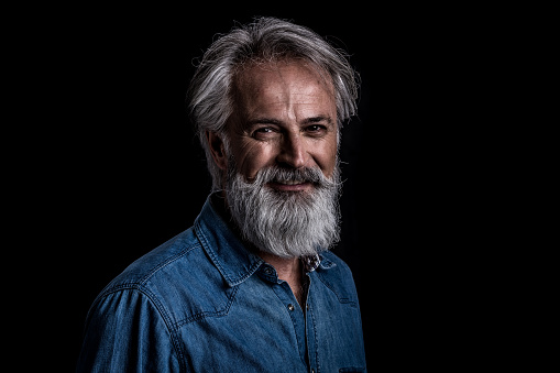 Smiling happy mature adult Caucasian guy. Confident man in blue jeans shirt against black background.