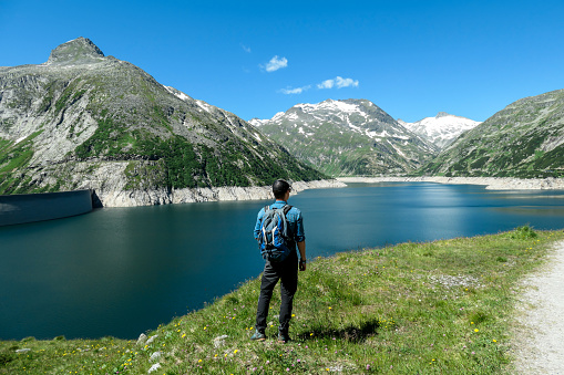 Man standing at the side of a lake at K?lnbreinsperre dam in Austria. The lake has navy blue color. High Alps around. There is a glacier in the back. The meadows blossoming with wild flowers. Calmness