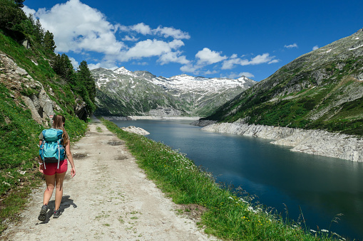 A woman wearing red shorts and hiking backpack hiking along the side of an artificial lake at a dam in Austria. The lake has navy blue color. High Alps around.  Adventure and discovering