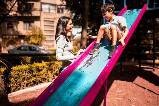 A young boy is on a slide, the mother is watching him. It's sunny outdoors, they are enjoying the weekend