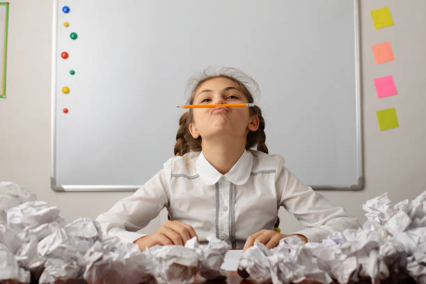 boring homework pictures Bored lazy young female student at the table with a lot of crumpled papers around her. Girl with two pigtails placing a pencil between her lips and nose. Concept of learning, boredom and laziness. boring homework twelve stock pictures, royalty-free photos & images
