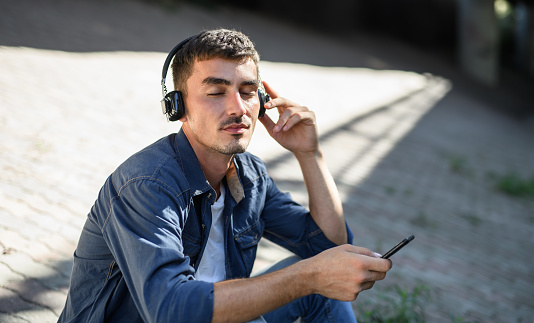 Portrait of young attractive man with headphones and smartphone sitting outdoors in city.