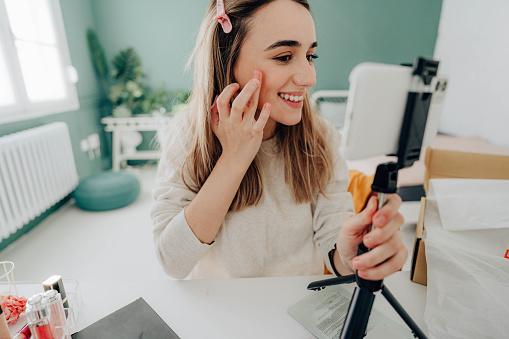 Photo of a young female blogger live streaming from her studio while applying, testing out beauty products, and reviewing them.