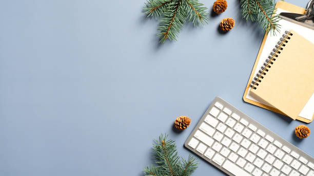 Cozy home desk table with keyboard, notebook, Christmas tree branches, cones on blue background. Top view, flat lay, copy space. Winter composition. Nordic hygge style concept stock photo