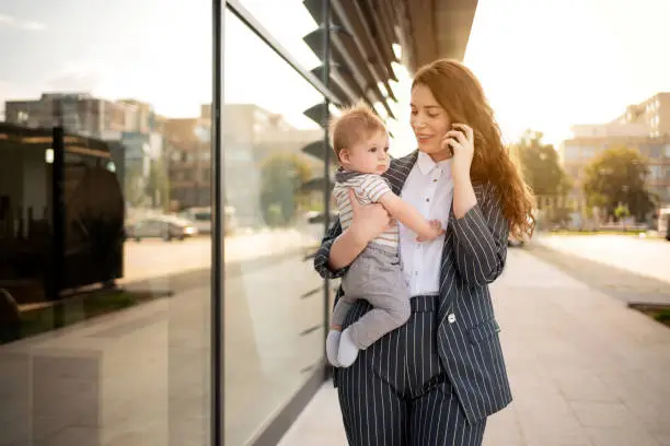 Photo of Businesswoman talking on her phone while walking on a sunny day and carrying her baby
