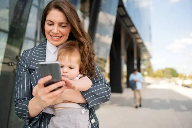 Photo of Young mother holding smart phone and texting while carrying her cute baby boy