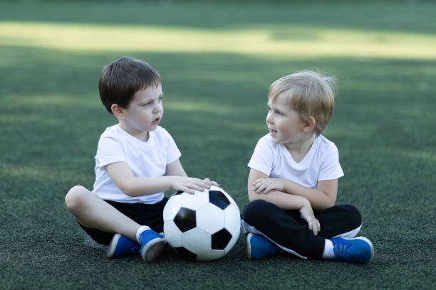 lifestyle of two children and soccer ball, sitting on grass, laughing and talking. concept about childhood, sports and communication of preschool. - ball horizontal outdoors childhood imagens e fotografias de stock