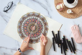 Woman drawing and coloring mandala in sketchbooks with colorful markers while having a breakfast