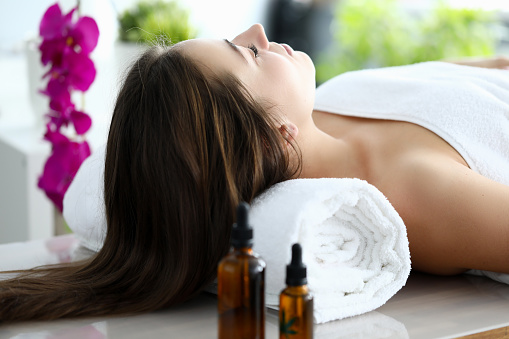 Woman with long hair lay on her back on massage table with her eyes closed. Spa client put his head on towel and wrapped white towel around his body. There are jars of aroma oils on table, flowers in background.