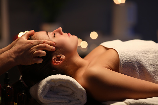 Woman lay on couch on her back with closed eyes and enjoy. Man make relaxing and therapeutic head massage at weight. Spa client has thrown her head back and rejuvenate. Wellness procedures in spa salon concept.