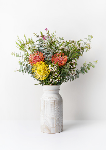 A beautiful floral arrangement of Australian native red and yellow Waratah flowers, eucalyptus leaves and wax flowers in a stylish white ceramic vase, with a white background.