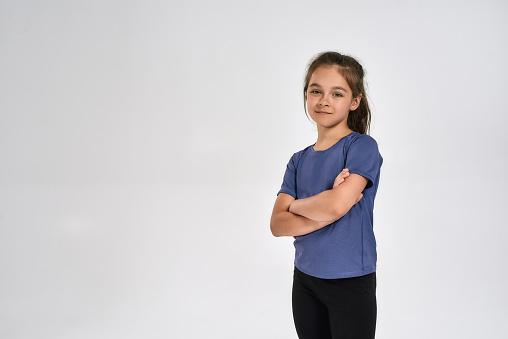 Little sportive girl child in sportswear looking at camera, while standing with arms crossed isolated over white background. Sport, training, fitness, active lifestyle concept. Horizontal shot