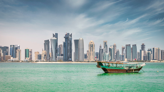 Modern Cityscape Panorama of the qatari Capital City of Doha. Waterfront Panorama at Doha Corniche Promenade with anchored typical qatari Dhow Cruise Boat for Touristic Doha Bay Tours. Modern business skyscraper Skyline of Doha in the background under blue summer sky. Doha, Qater, Middle East