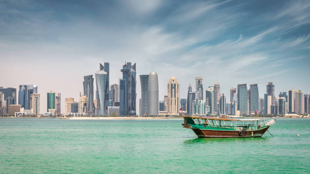 Doha Cityscape Qatar Panorama Qatari Dhow Doha Cruise Boat Qatar Middle East Modern Cityscape Panorama of the qatari Capital City of Doha. Waterfront Panorama at Doha Corniche Promenade with anchored typical qatari Dhow Cruise Boat for Touristic Doha Bay Tours. Modern business skyscraper Skyline of Doha in the background under blue summer sky. Doha, Qater, Middle East dhow photos stock pictures, royalty-free photos & images
