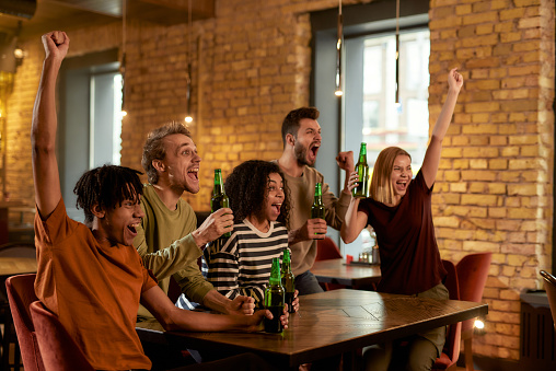 Happy friends in the bar watching sports match on TV together, drinking beer and cheering for team. People, leisure, friendship and entertainment. Selective focus. Horizontal shot