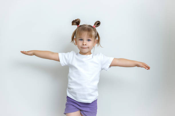 une fille mignonne de 4 ans dans un t-shirt blanc se tient avec ses bras tendus aux côtés sur un fond léger. - child caucasian little girls 3 4 years photos et images de collection