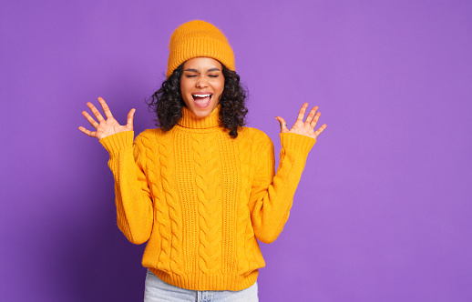 Delighted young ethnic woman in knitted yellow sweater and hat  gesticulating and screaming with closed eyes while having fun against purple background