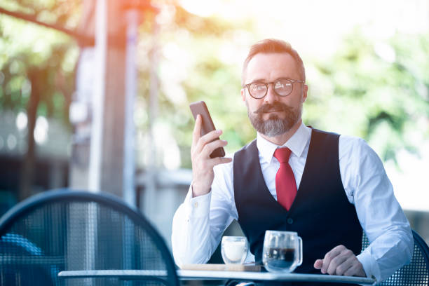 homme d’affaires d’âge moyen s’asseyant à un café buvant le café et utilisant le téléphone intelligent. mand caucasien de hansome retenant le téléphone portable souriant à l’appareil-photo au café. - coffee shop coffee break coffee cup holding photos et images de collection