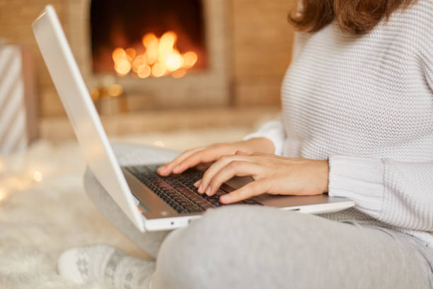 istock Faceless woman typing message on her laptop computer as she relaxes near burning warm fire, unknown female working online while sitting on floor with crossed legs, holding hotebook on her knees. 1288406992