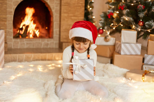 istock Cute little child in red festive hat with Christmas gift box posing at home while sitting on floor on soft carpet near fireplace and xmas tree. Little charming girl with pigtails holds gift box. 1288408036