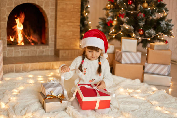 istock Cute female kid with pigtails sitting on floor and pulling ribbon from her present box, opening her gift, wearing pullover and santa hat, posing with fireplace and Christmas tree on background. 1288408045