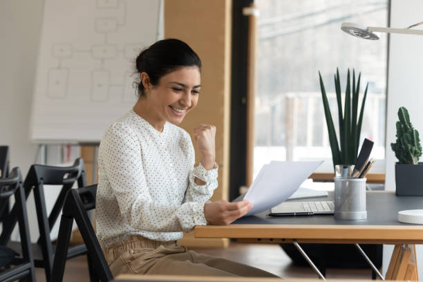 Happy millennial indian woman reading paper letter with good news. Happy millennial indian ethnic woman reading paper letter with amazing good news, making yes i did it gesture, celebrating banking loan approval, passed professional qualification test in office. personal loan stock pictures, royalty-free photos & images