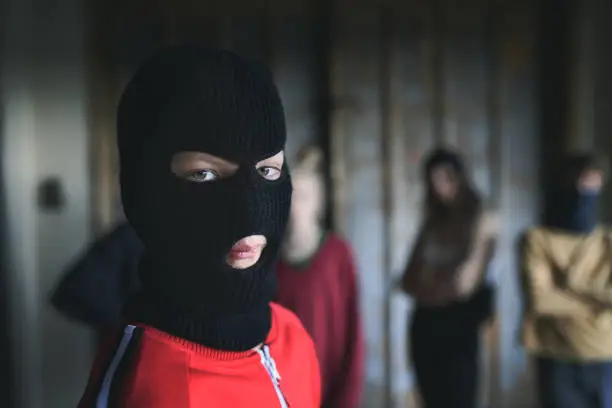Boy with mask and teenagers gang standing indoors in abandoned building, looking at camera.