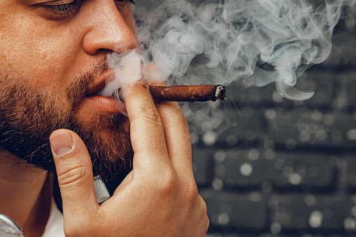 Bearded man with stylish haircut in profile holding electronic cigarette in hand and exhaling smoke in air, black and white waist-up shot