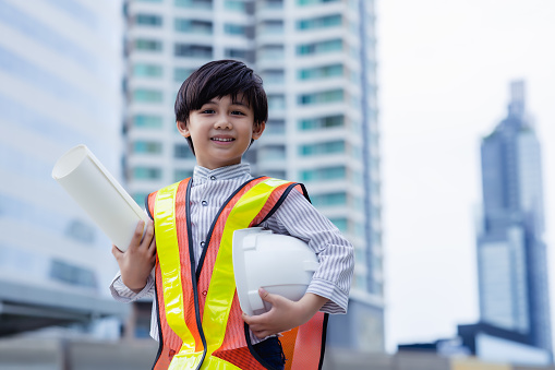 Handsome little engineer asian boy holding safety helmet and blueprint. Adorable little smart kid wear protective workwear and look at city with smile face and happy Dream kid and future life concept