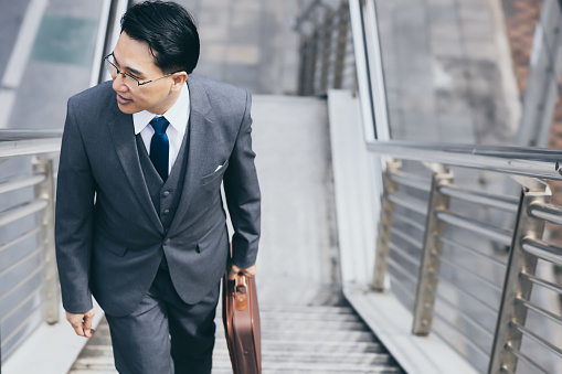 Happy businessman going to his office for work in morning by walking on steps of overpass with happy and smile face Handsome man wear suit, necktie and holding briefcase Business and finance concept