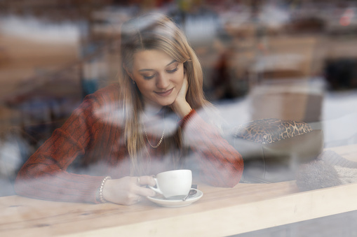 A young pretty girl with a cup of coffee in a cafe in front of the window. View from outside through glass with reflections.