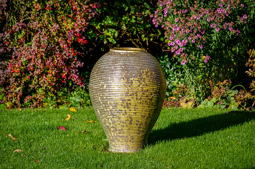 A standalone image of a single, large green pot in the shape of an amphora with a backdrop of a typical mixed flower bed in an English Residential Garden on a late autumn morning in 2020. The mottled, ridged green glaze on the pot blends perfectly with the green grass of the lawn on which it sits and the dark maroon colour of the Japanese Barberry shrub, behind and to its left, coupled with the brighter green foliage and pink/mauve flowers of the Chrysanthemum, behind and to its right, all bathed in low level autumn sunlight complete this atmospheric image.