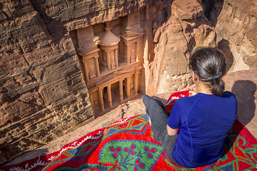 Woman traveler sitting on carpet viewpoint in Petra ancient city looking at the Treasury or Al-khazneh, famous travel destination of Jordan and one of seven wonders of the modern world. UNESCO World Heritage site