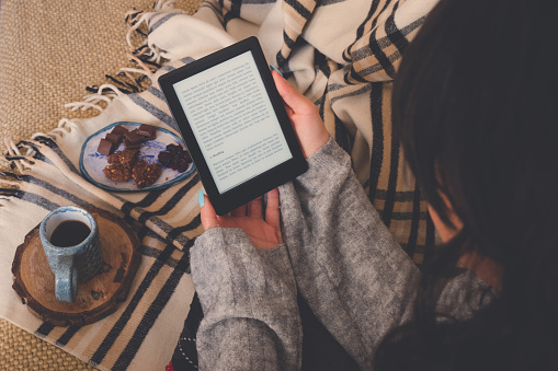 Top view of caucasian woman reading an e-book in the e-book reader with a cup of coffee and cocoa cookies. Cozy winter day at home and hygge concept. The text on the e-book reader is an \