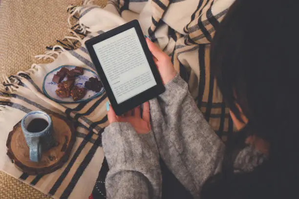 Top view of caucasian woman reading an e-book in the e-book reader with a cup of coffee and cocoa cookies. Cozy winter day at home and hygge concept. The text on the e-book reader is an "Lorem Ipsum"