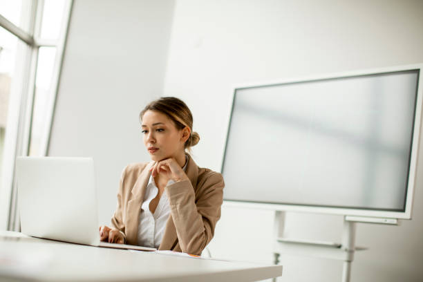 istock Young woman working on laptop in bright office with big screen behind her 1288459457