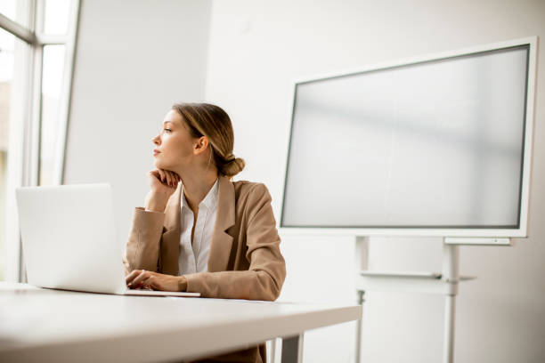 istock Young woman working on laptop in bright office with big screen behind her 1288459471