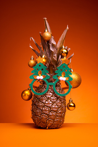 Pineapple in gold color decorated with glitter baubles, isolated on orange background wearing funky green x-mass sunglasses in the shape of a tree. Alternative Christmas tree for tropical December holidays.