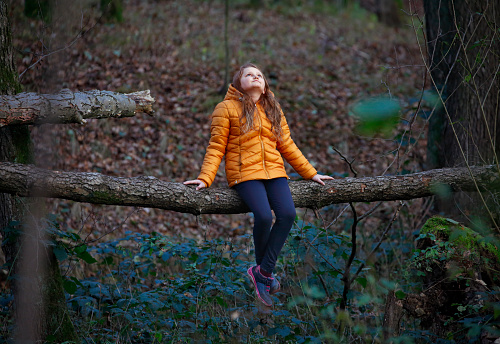 Young pensive lonely girl sitting on the tree trunk in the autumn forest