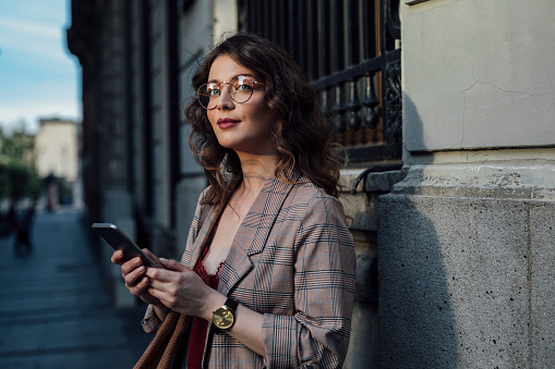 A young, smart-looking, elegant woman standing in the street, holding her phone.