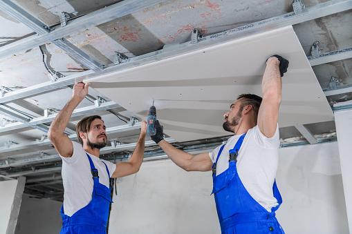 The adjuster holds the plasterboard firmly while a colleague fixes it to the ceiling with a drill