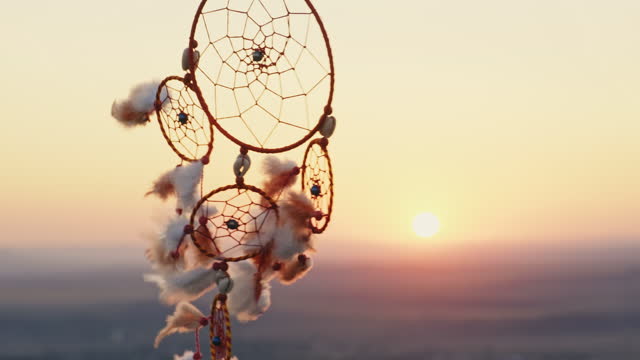 Dreamcatcher with orange feathers rotate in mountains, swaying with light wind slow motion agains blue sky at sunset in summer. Amulet boho style, Indian style. Bright disk of sun, rays of sun